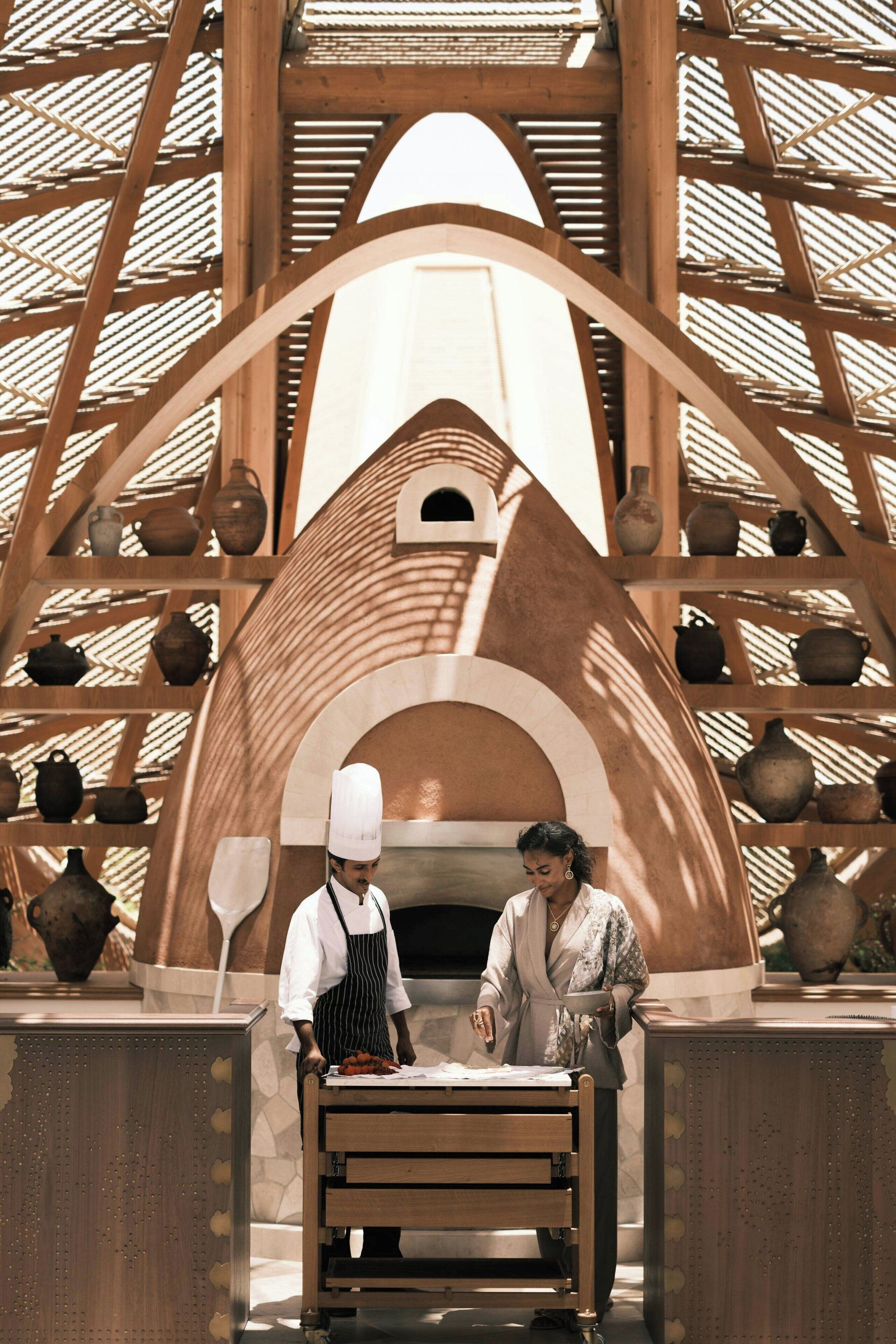 A chef and a woman standing in front of a traditional oven with pottery on shelves.