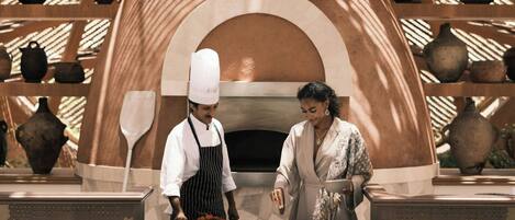 A chef and a woman standing in front of a traditional oven with pottery on shelves.