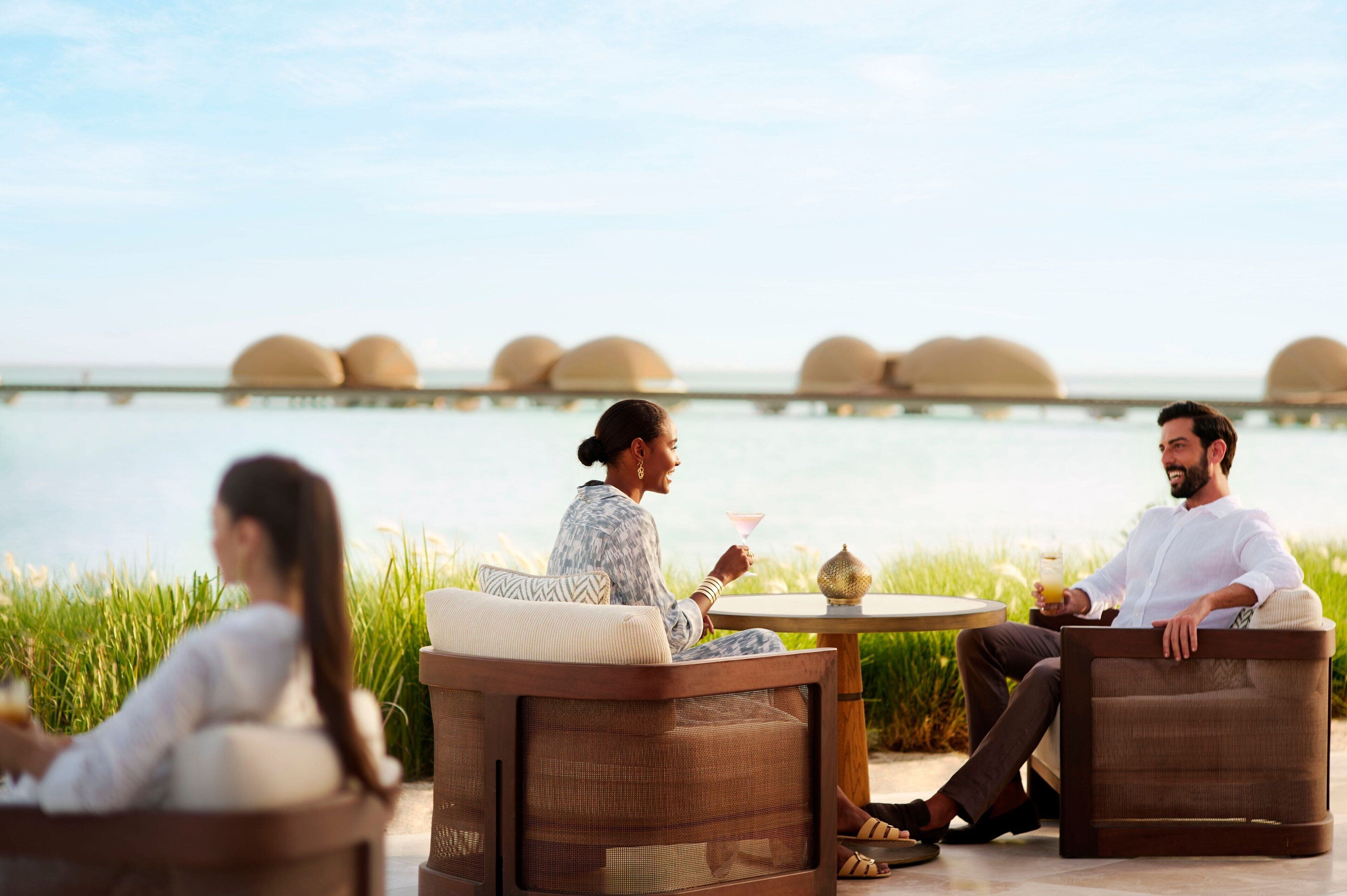 Una pareja y una mujer sentados en una mesa con vista a un cuerpo de agua y bungalows sobre el agua al fondo.
