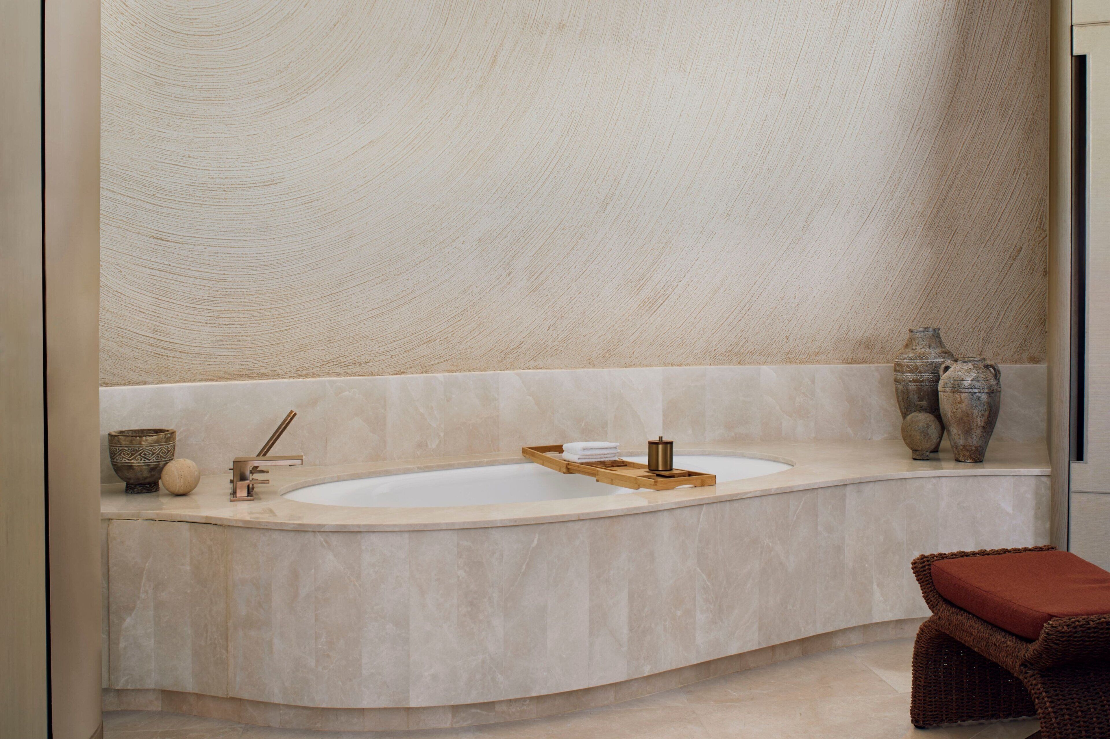 Ein modernes Badezimmer mit einer großzügigen, gebogenen Badewanne, zwei Waschbecken und einem Holztablett. An den Wänden befinden sich zwei große Vasen und ein Korbstuhl mit Kissen.