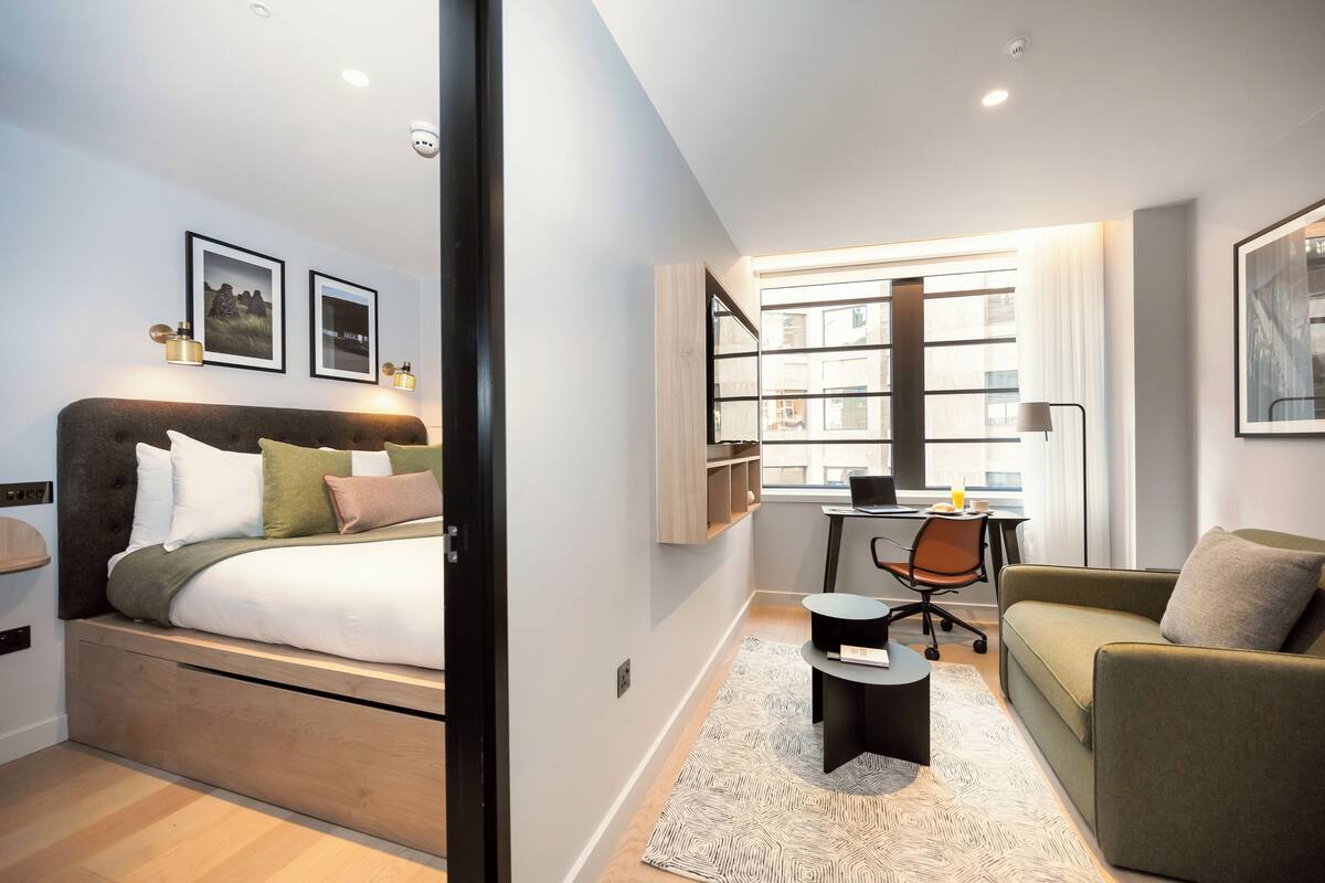 A modern hotel room with a bed, a sofa, a desk, and a window with blinds.