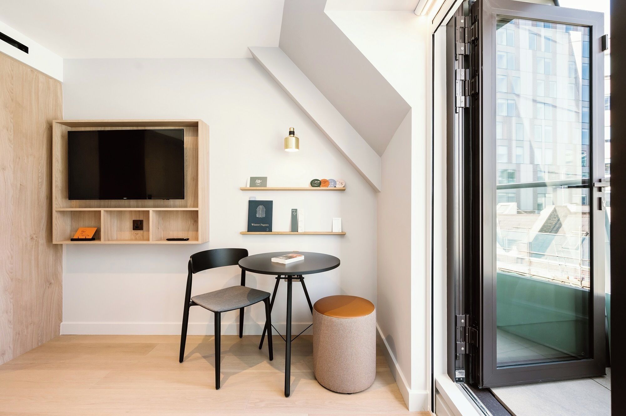 Een moderne, compacte leefruimte met een flatscreen televisie bevestigd aan een houten paneel, een klein rond tafeltje met twee stoelen en een rond, geweven krukje. Daarboven hangen twee zwevende planken met boeken en decoratieve objecten. Een groot raam met een glazen deur is zichtbaar, met uitzicht op de stad.