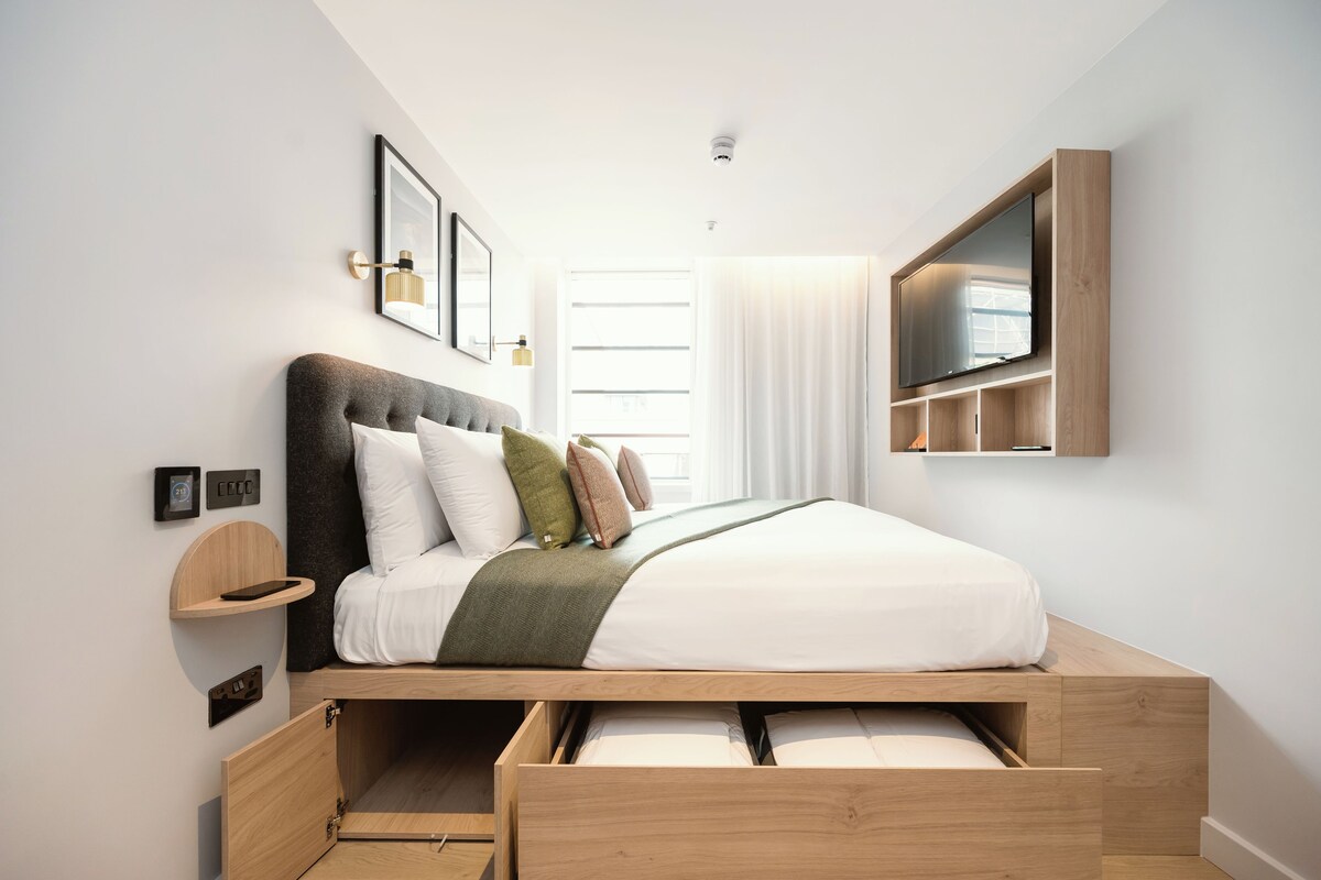 A modern bedroom with a wooden bed frame, a built-in TV, and a bedside table.