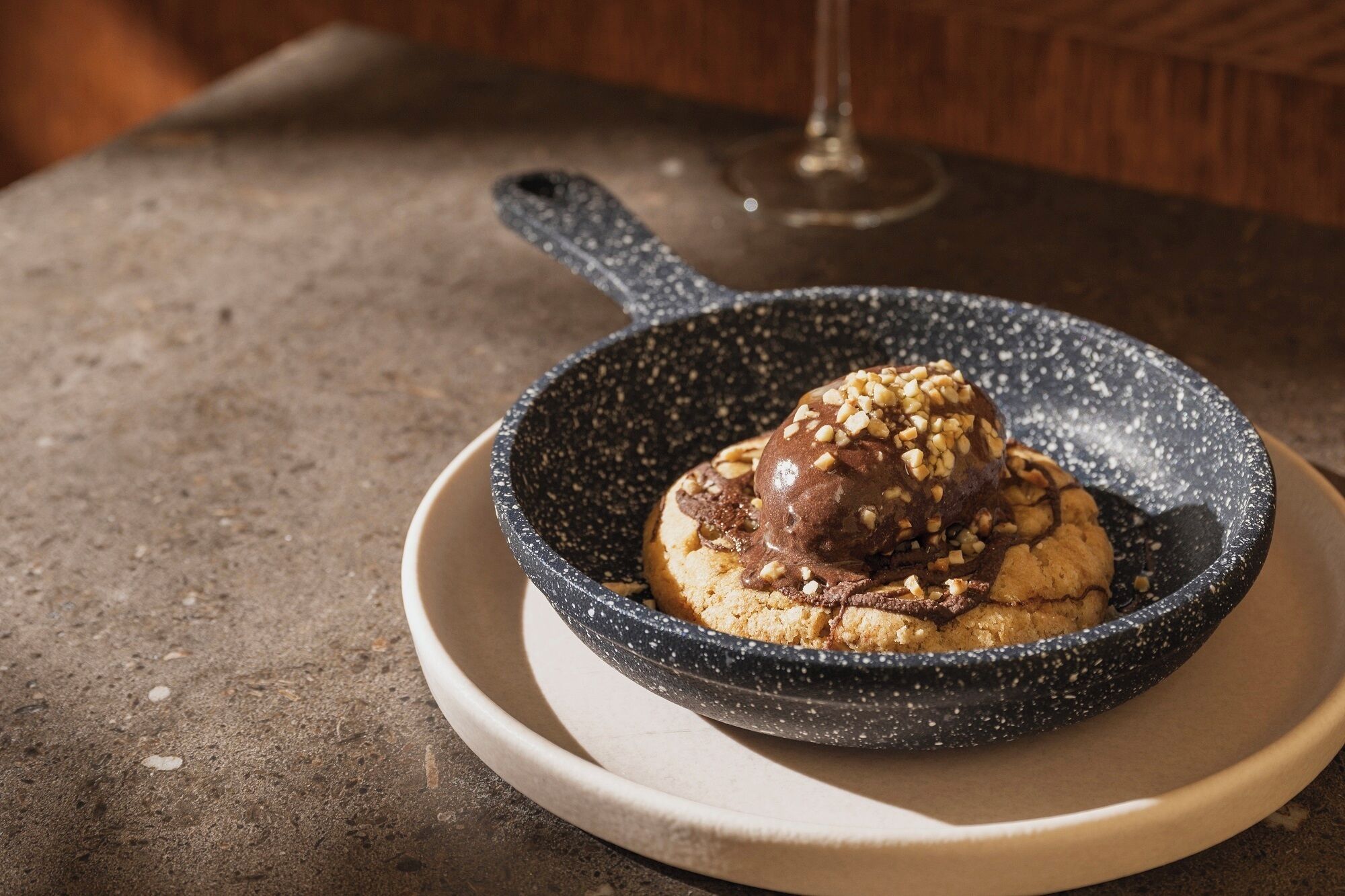 Un postre en un tazón moteado sobre un plato, con una copa de vino al fondo.