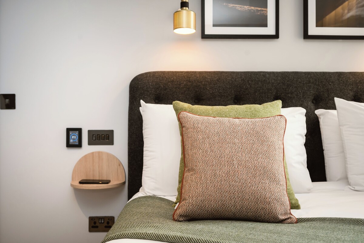 A neatly arranged hotel room with a bed, a bedside table, a wall-mounted shelf, and a wall-mounted light fixture.