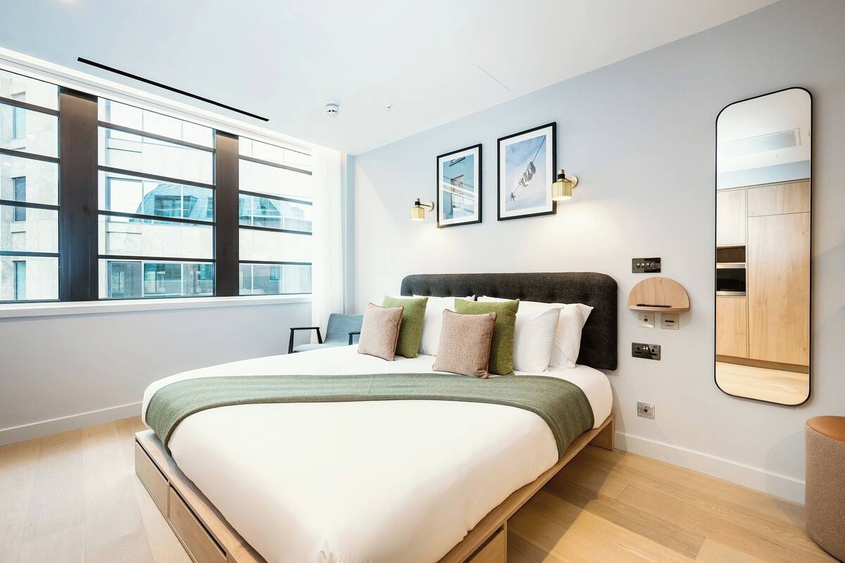 A modern bedroom with a large bed, a window with blinds, a wall-mounted mirror, and framed pictures.