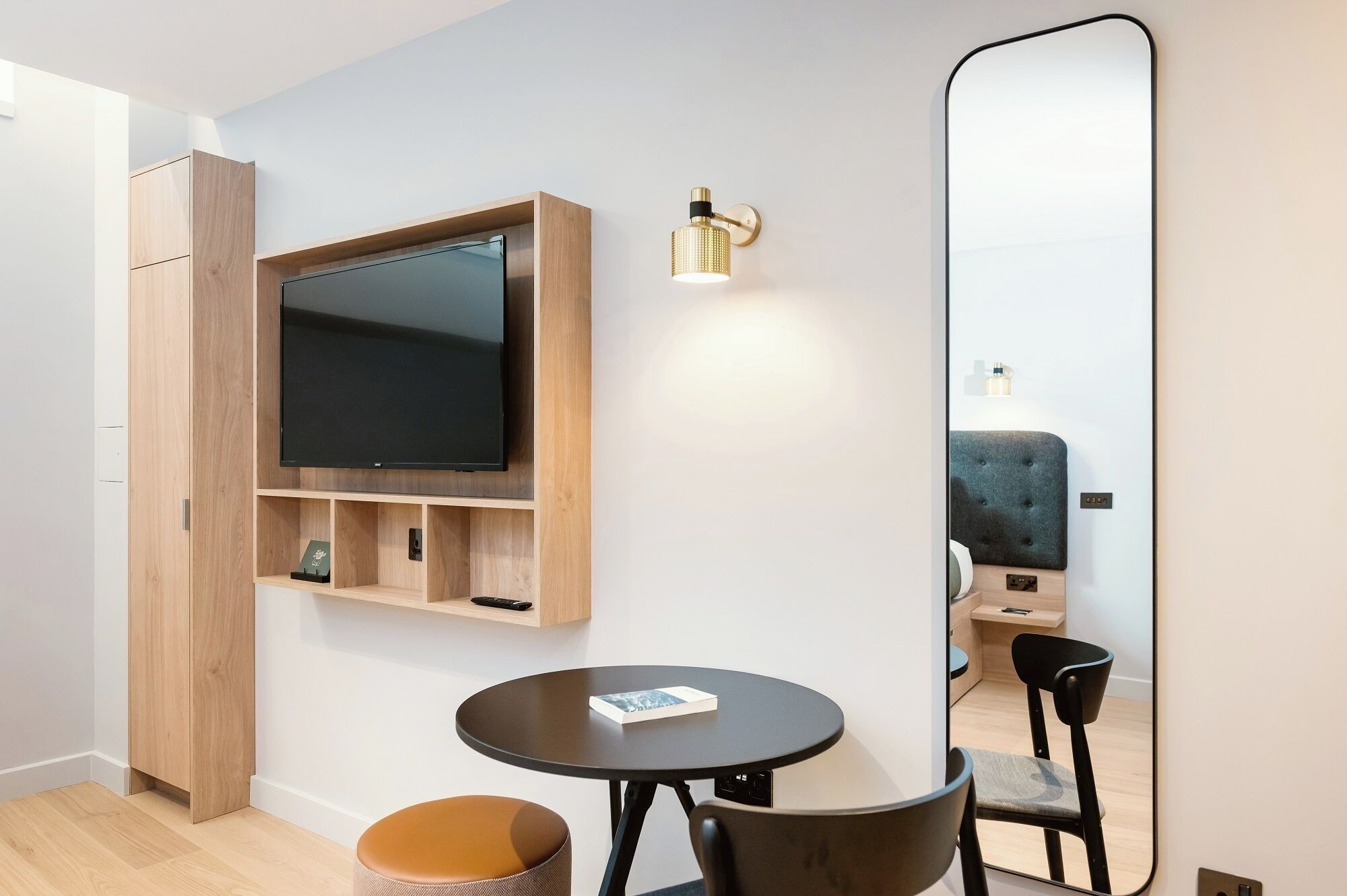 Een moderne kamer met een flatscreen televisie bevestigd aan een houten plank, een ronde tafel met een stoel, een grote spiegel en een klein voetenbankje.