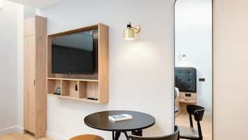 Een moderne kamer met een flatscreen televisie bevestigd aan een houten plank, een ronde tafel met een stoel, een grote spiegel en een klein voetenbankje.