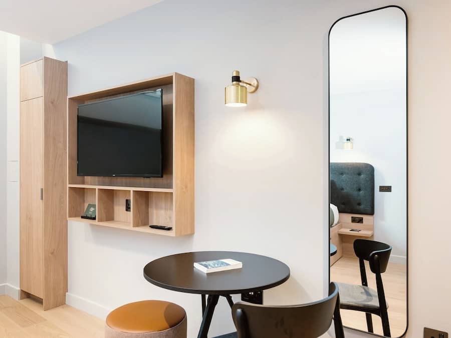 Een moderne kamer met een flatscreen televisie bevestigd aan een houten plank, een ronde tafel met een stoel, een grote spiegel en een klein voetenbankje.
