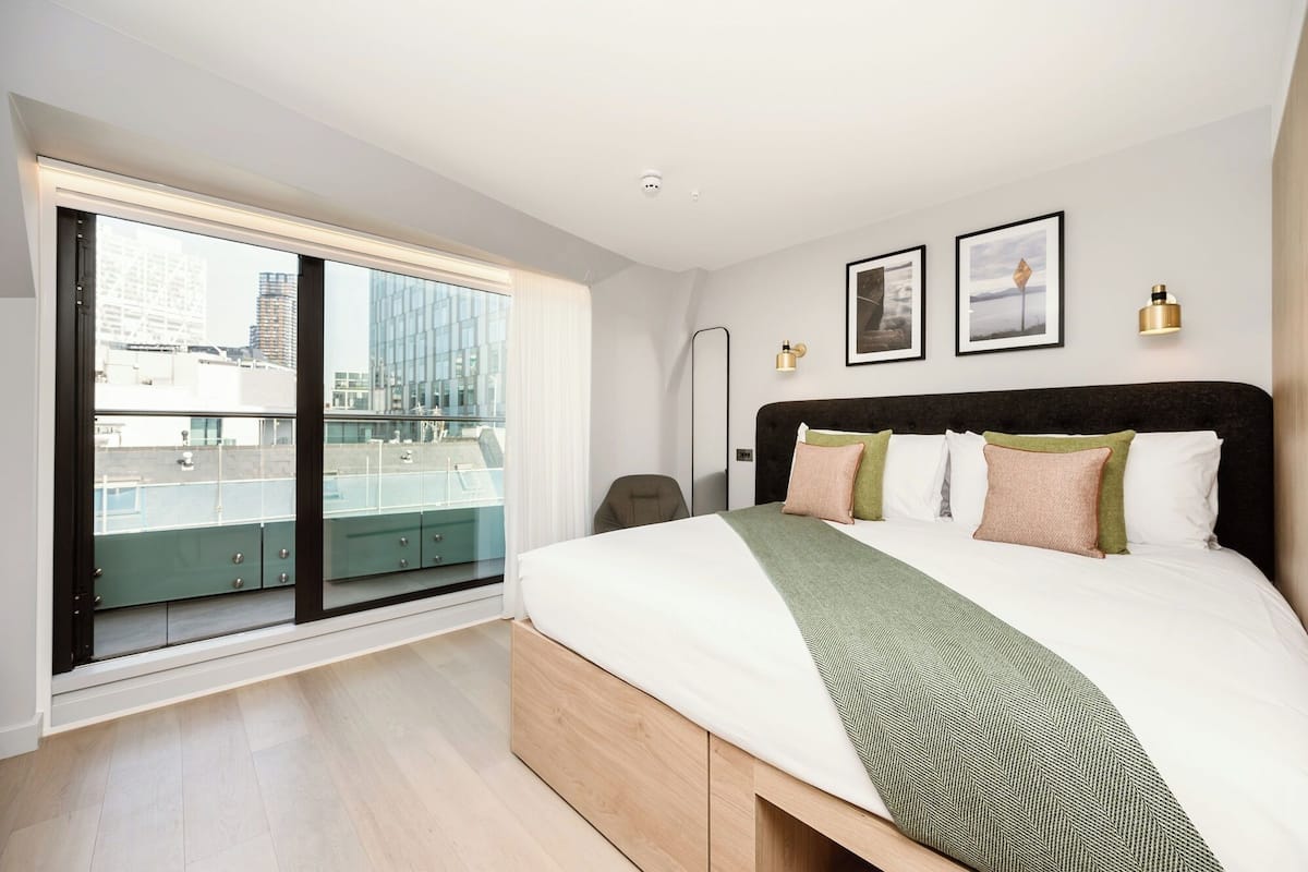 A modern hotel room with a large bed, a view of the city through floor-to-ceiling windows, and a balcony with a green railing.
