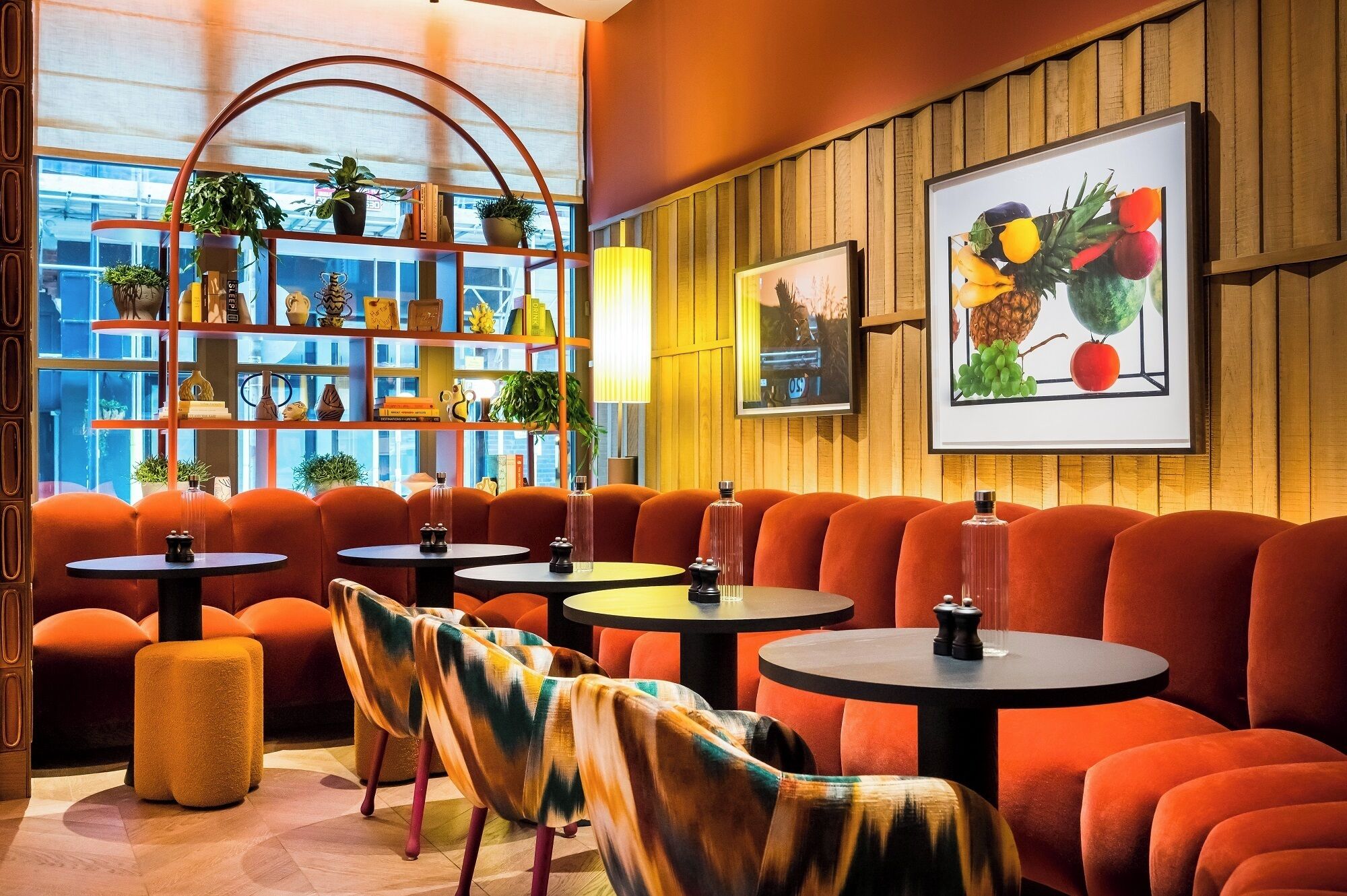 A modern interior with orange seating, round tables, and a bookshelf with plants.