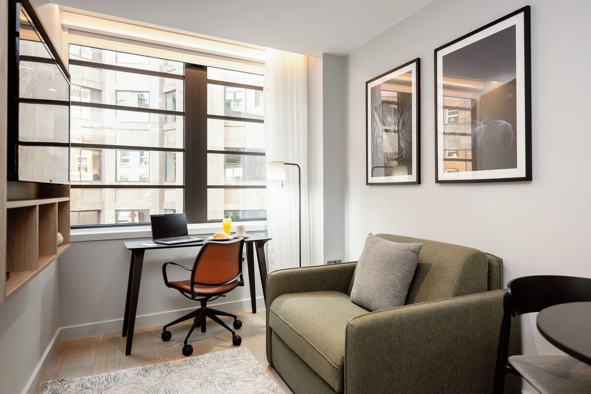 A modern room with a desk, chair, and sofa. There is a window with a view of buildings outside.