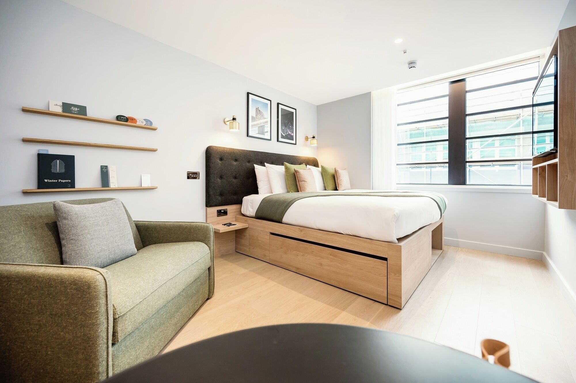 A modern bedroom with a bed, a green sofa, wooden shelves, and a large window.