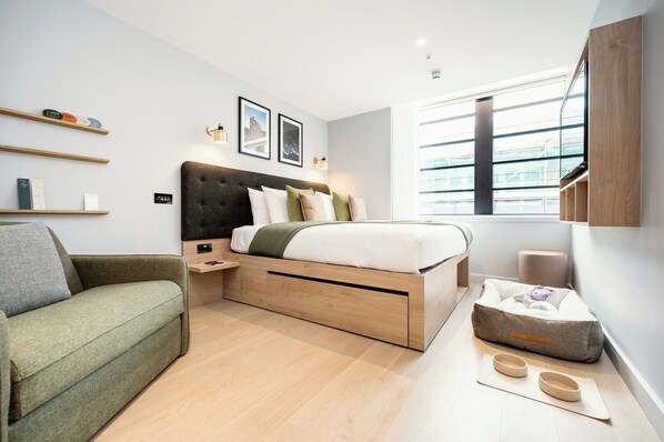 A modern bedroom with a bed, bedside tables, a green sofa, a window with blinds, and a small wooden cabinet. - Wilde Aparthotels London Liverpool St. (London)