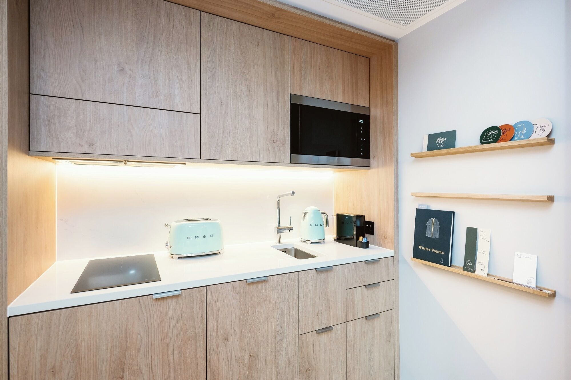 A modern kitchen with wooden cabinets, a microwave, a toaster, and a coffee machine. There are shelves with decorative items and a book titled "Winter Papers".