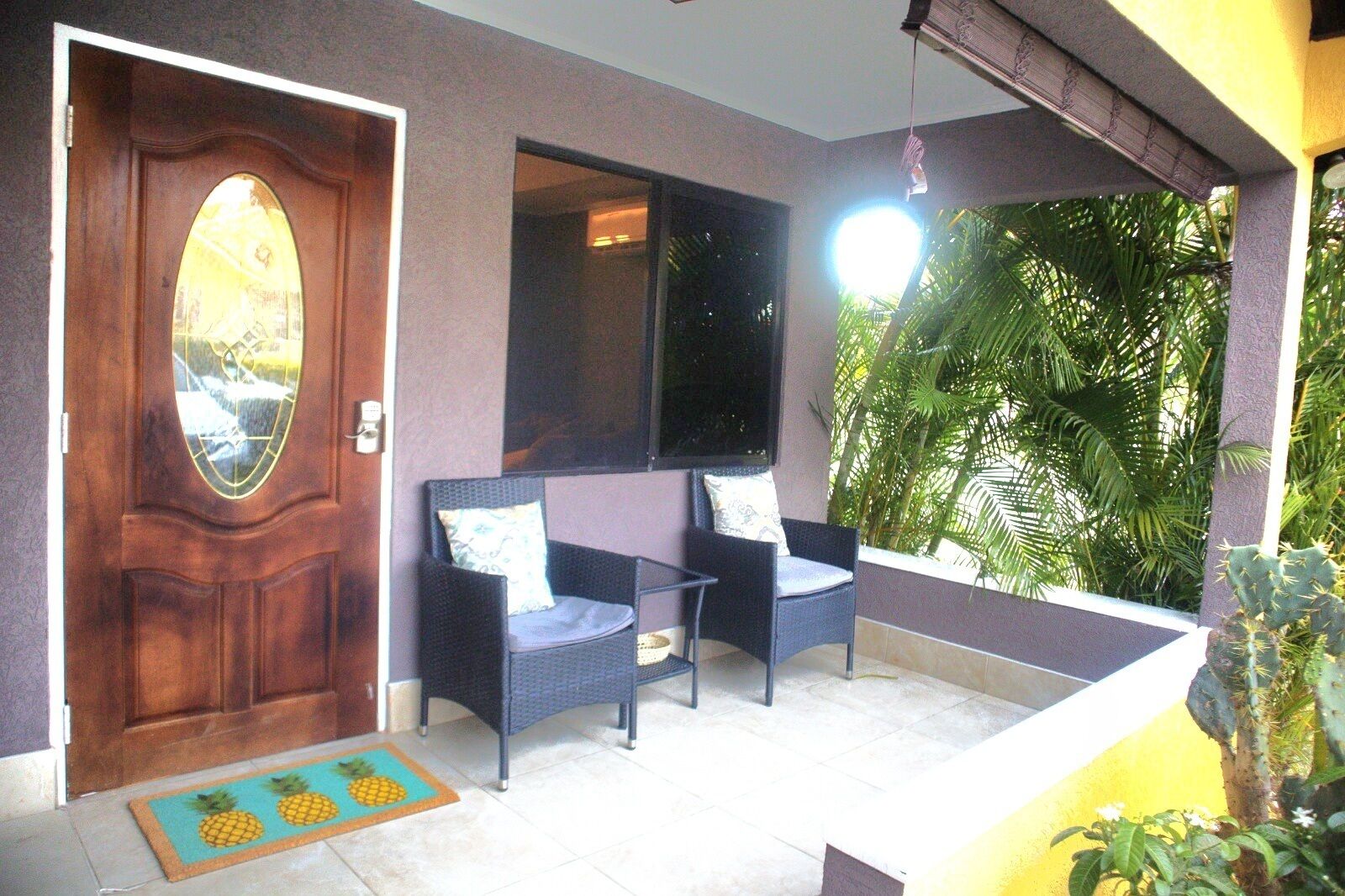 A room with a wooden door, two wicker chairs, a large window, and a view of greenery.