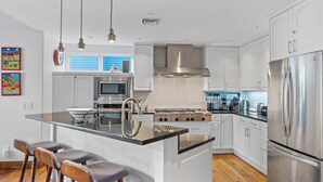 Cocina moderna con armarios blancos, electrodomésticos de acero inoxidable, una isla central con taburetes altos, iluminación colgante y una ventana con persianas azules.