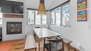 Un comedor con una mesa larga, sillas, una lámpara colgante, chimenea y ventanas amplias con vistas a edificios.