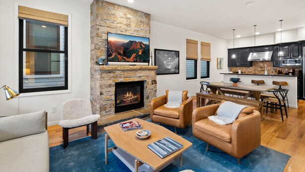 A modern living room with a stone fireplace, leather sofas, a wooden coffee table, and a dining area with a kitchen in the background.