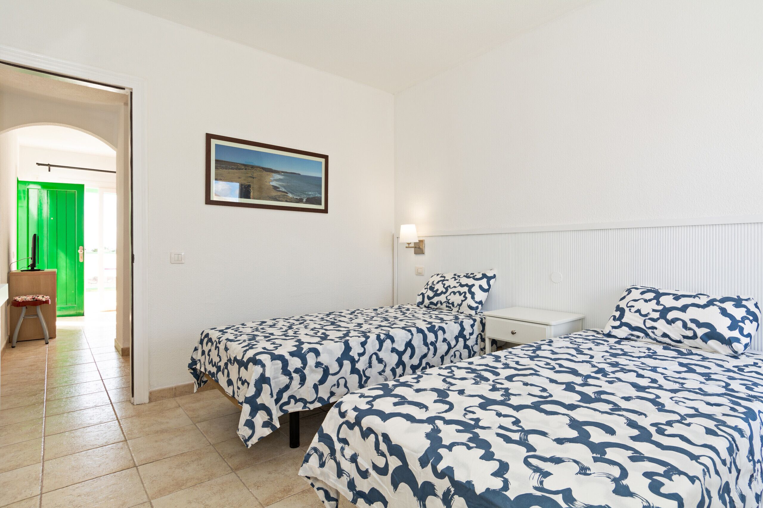 Habitación de hotel con dos camas, una puerta verde, un cuadro enmarcado en la pared y una mesita de noche blanca.