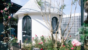 A white building with two black doors, surrounded by a garden with pink flowers and a tree. - Trulli Villa Homestay (Don Duong)