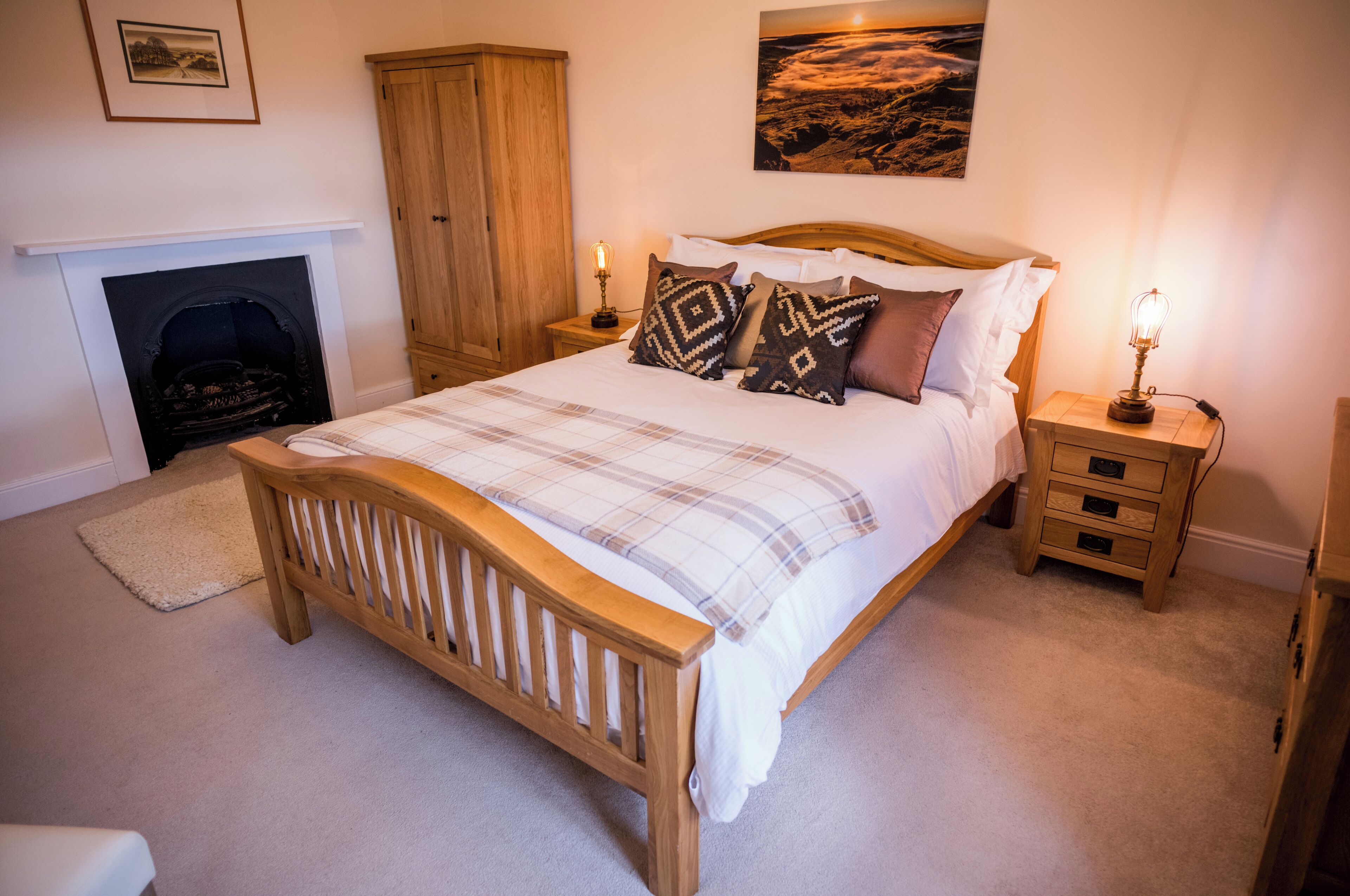 A bedroom with a wooden bed, a fireplace, a nightstand with a lamp, and a painting on the wall.