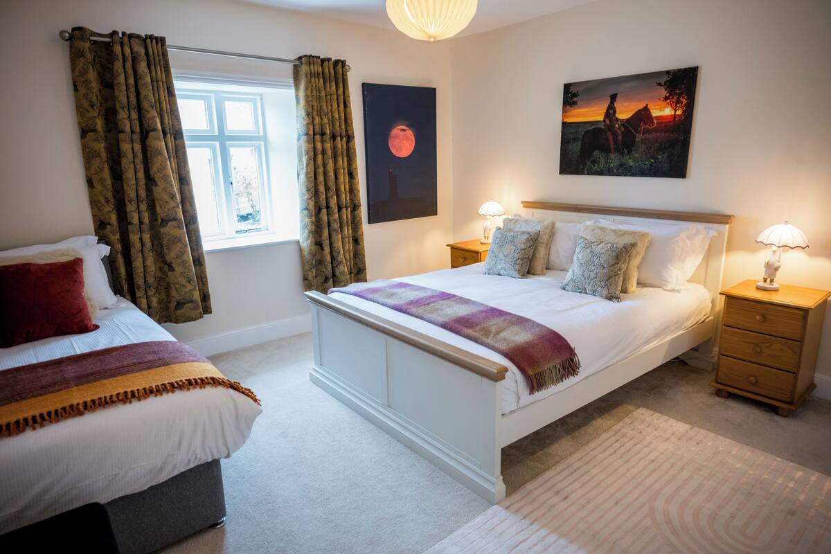 A bedroom with a large bed, two bedside tables, a nightstand, a window with curtains, and a wall-mounted picture.