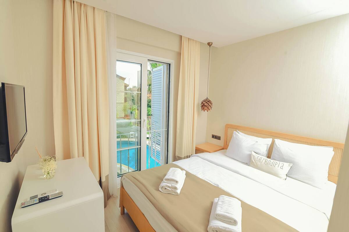 A hotel room with a bed, a TV on a side table, a balcony with curtains, and a view of the outdoors.