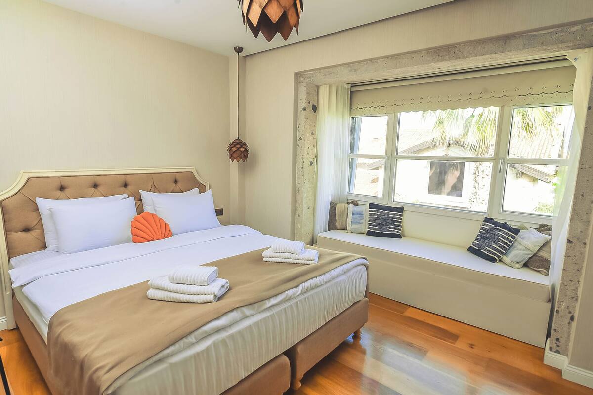 A neatly made bed with a brown headboard, a window seat with cushions, and a hanging pendant light.