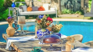 Une table à manger installée à l’extérieur, avec des chaises en osier, une piscine en arrière-plan et différentes décorations de table, notamment un vase avec des fleurs et des bougies.