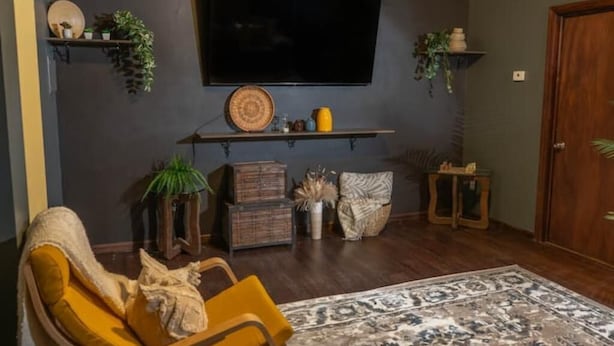 Un salon doté d’un téléviseur à écran plat fixé au mur, d’un fauteuil jaune, d’un tapis à motifs et de divers objets décoratifs disposés sur des étagères et des tables d’appoint.