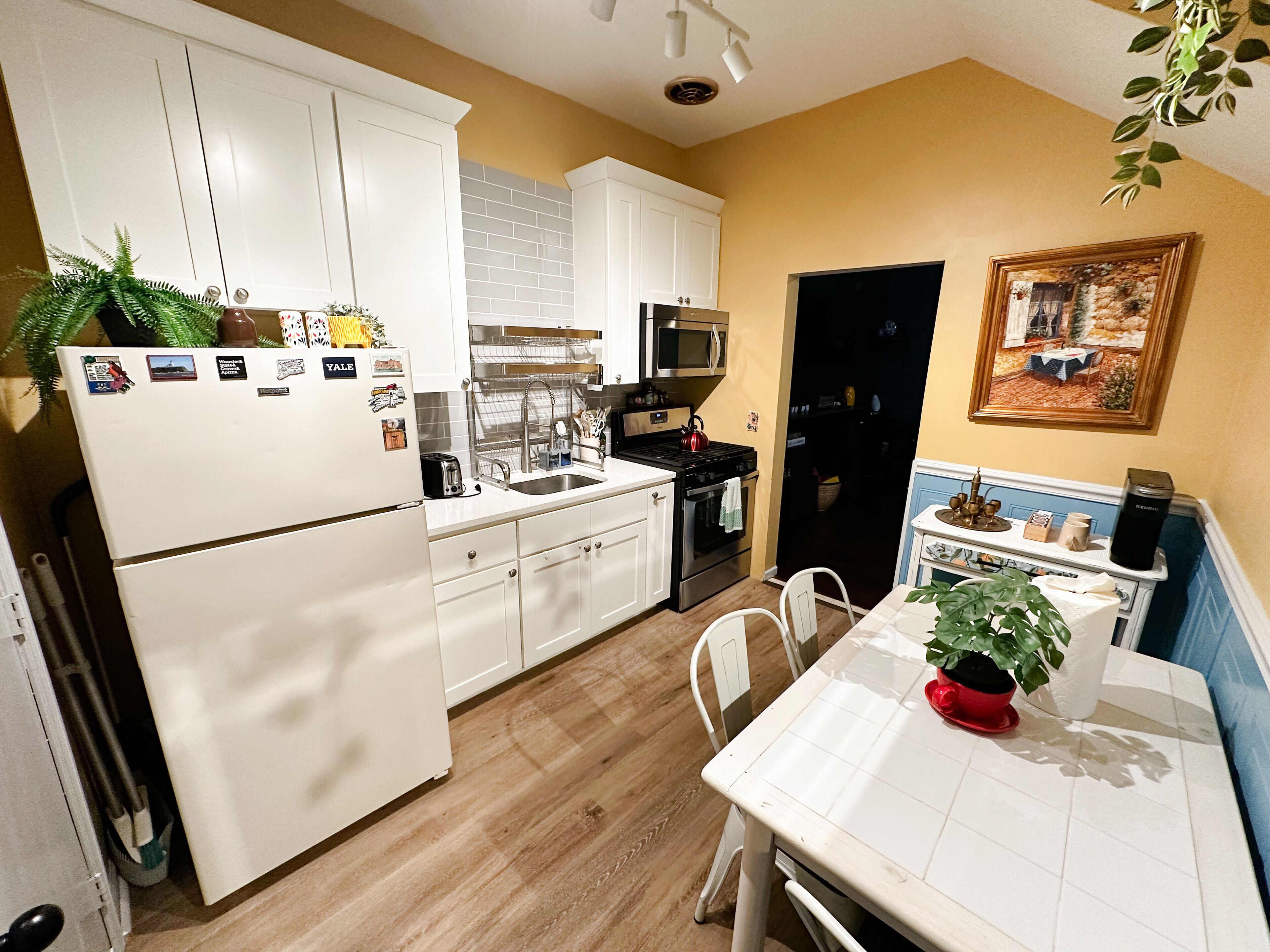 A kitchen with white cabinets, a stainless steel stove, and a microwave. There is a dining table with chairs and a potted plant.