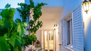 A narrow outdoor corridor with potted plants, a door leading to an interior space, and a wall-mounted light fixture. - Three Bedroom Apartment 7-9 Adults First Floor (Paros)