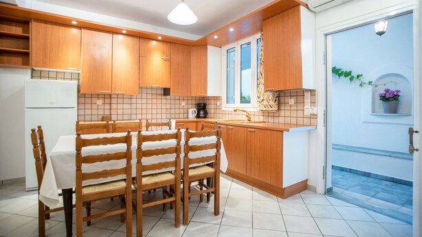 A modern kitchen with wooden cabinets, a tiled backsplash, a dining table with chairs, and a window with a view. - Three Bedroom Apartment 7-9 Adults First Floor (Paros)