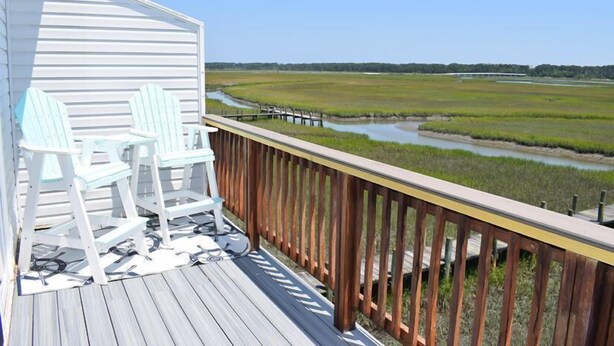 A wooden deck with two white Adirondack chairs overlooking a marshy landscape with a small water body and a wooden dock.