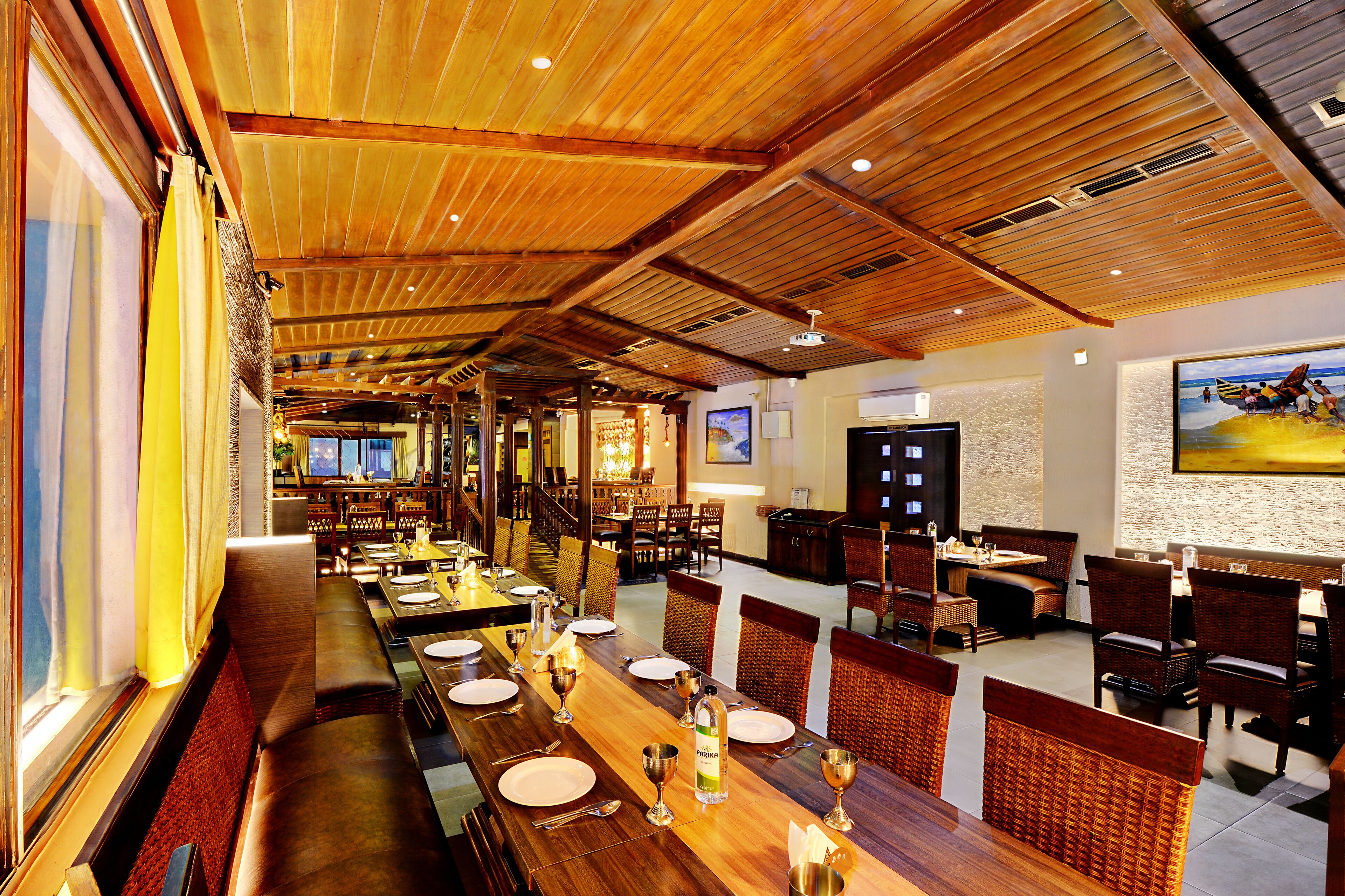 Um restaurante amplo com vigas de madeira no teto, longas mesas de jantar e cadeiras de vime.
