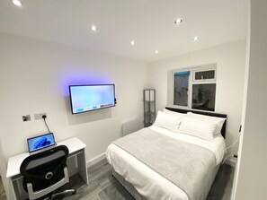 A modern bedroom with a bed, a desk with a laptop, a TV mounted on the wall, and a window with blinds.