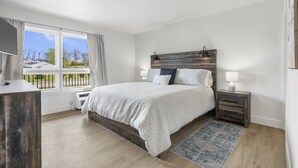 A bedroom with a large bed, a nightstand with a lamp, a window with curtains, and a view of a house and trees.