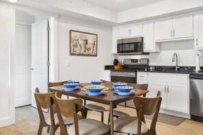 A modern kitchen with white cabinets, a microwave, an oven, and a sink. There is a dining table with blue and white bowls and a painting on the wall.