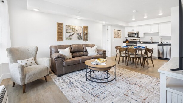 A modern living room with a brown leather sofa, a beige armchair, and a coffee table. A kitchen with white cabinets and stainless steel appliances is visible in the background.