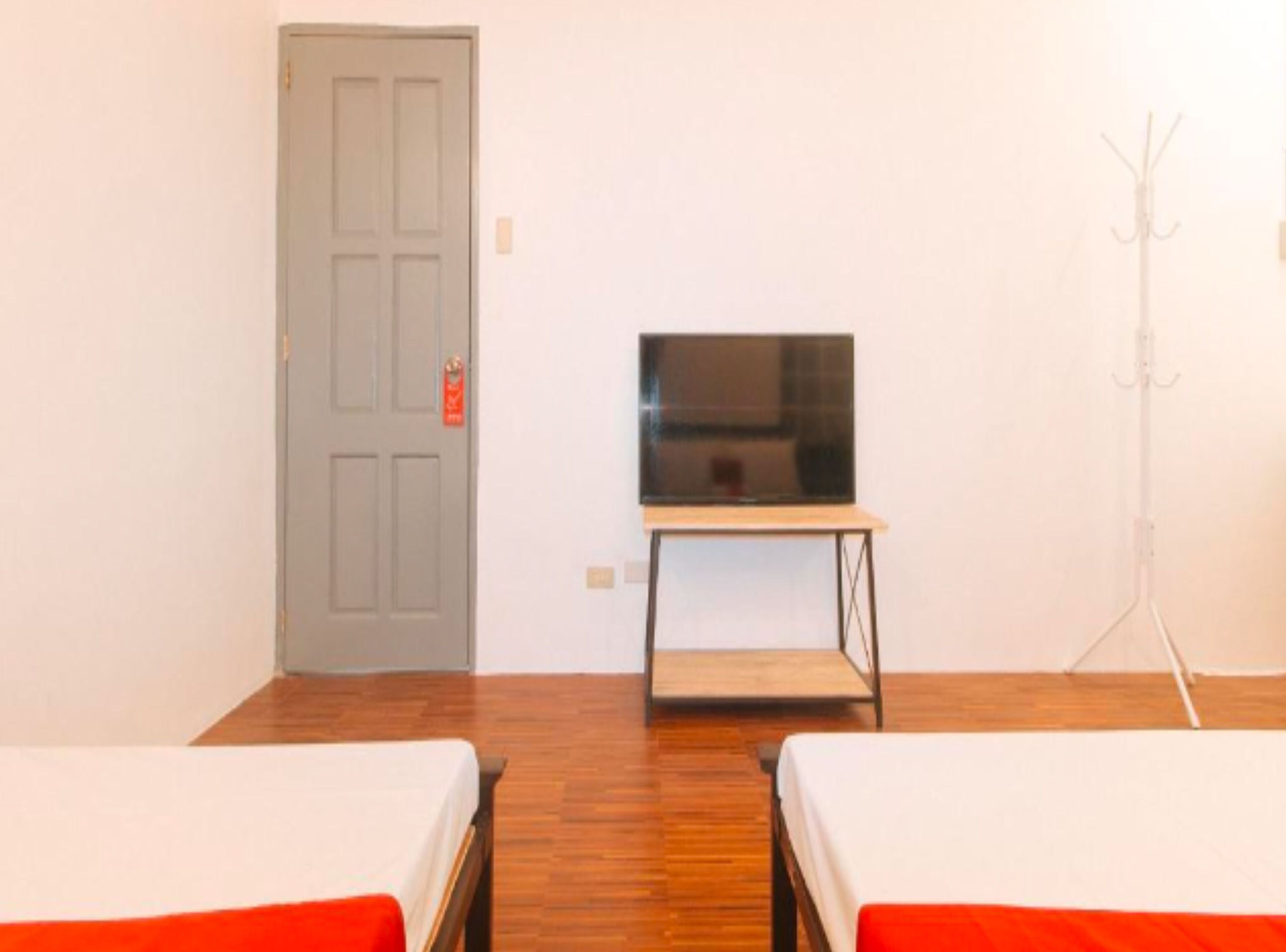 A room with two beds, a television on a stand, a coat rack, and a door with a red sign.