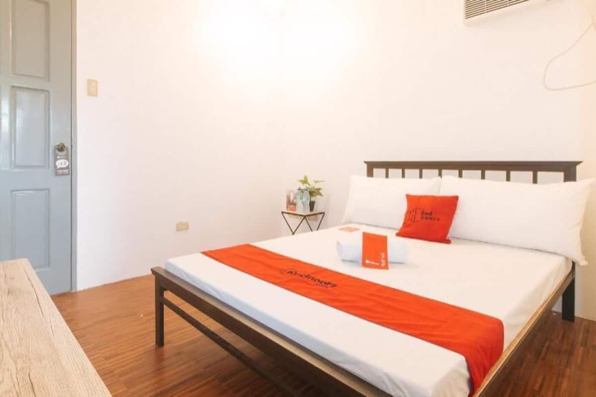 Una habitación de hotel con una cama, una mesita de noche con lámpara, una planta pequeña y una puerta.