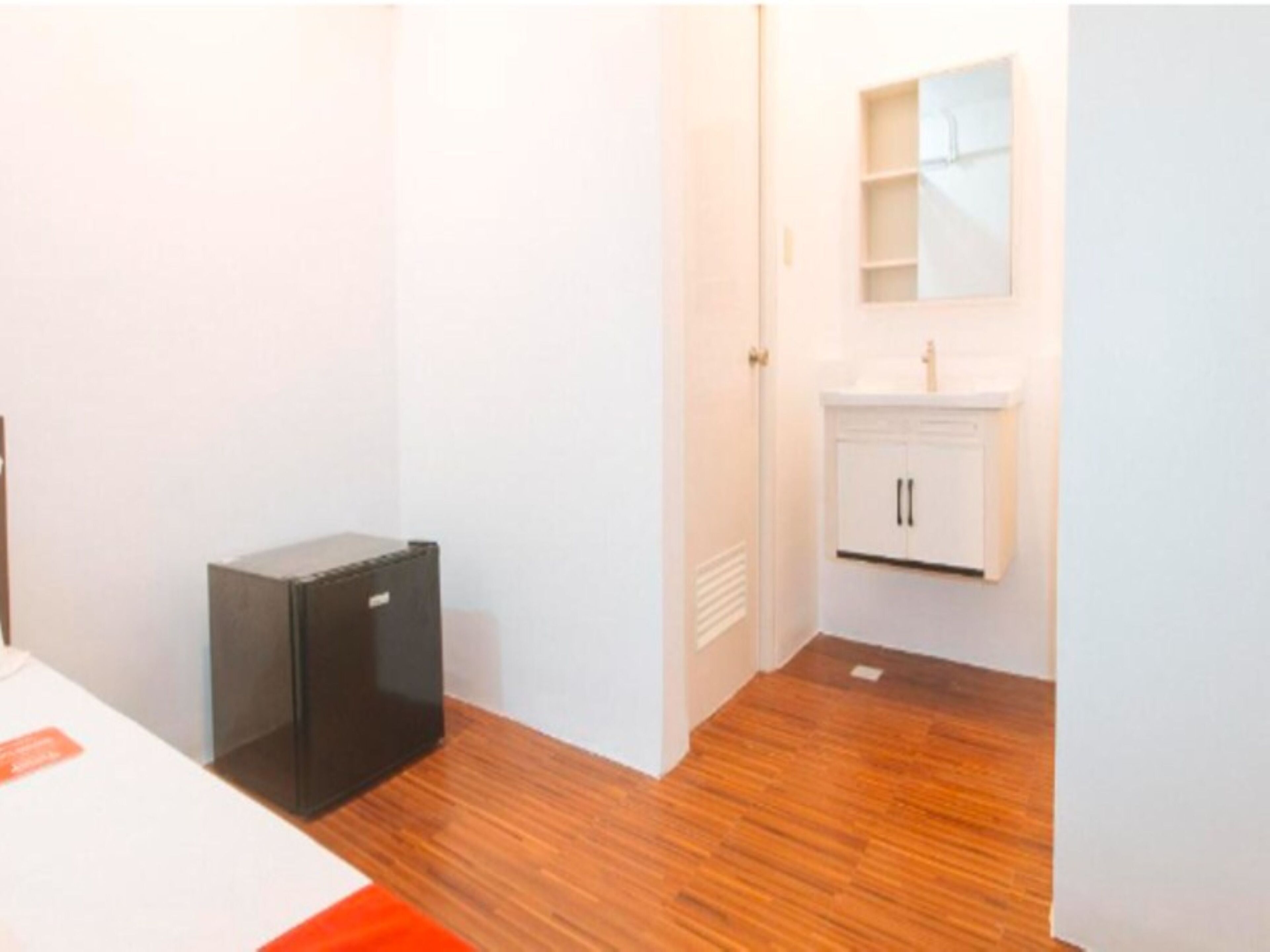 A small room with a wooden floor, a white wall, and a white cabinet with a mirror above it.