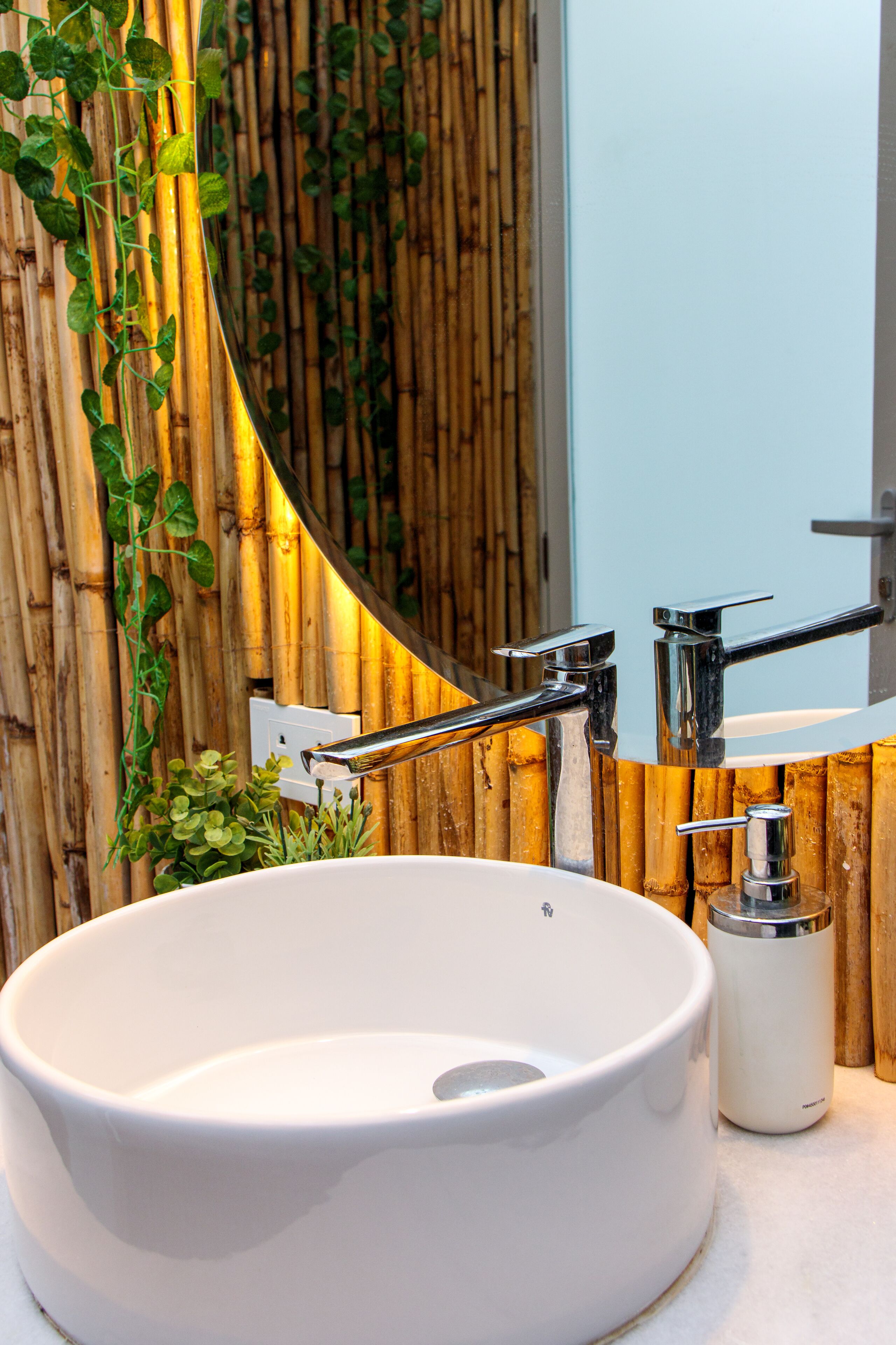 Un baño con un lavamanos redondo blanco, grifos cromados, un espejo y una pared de bambú con plantas verdes.