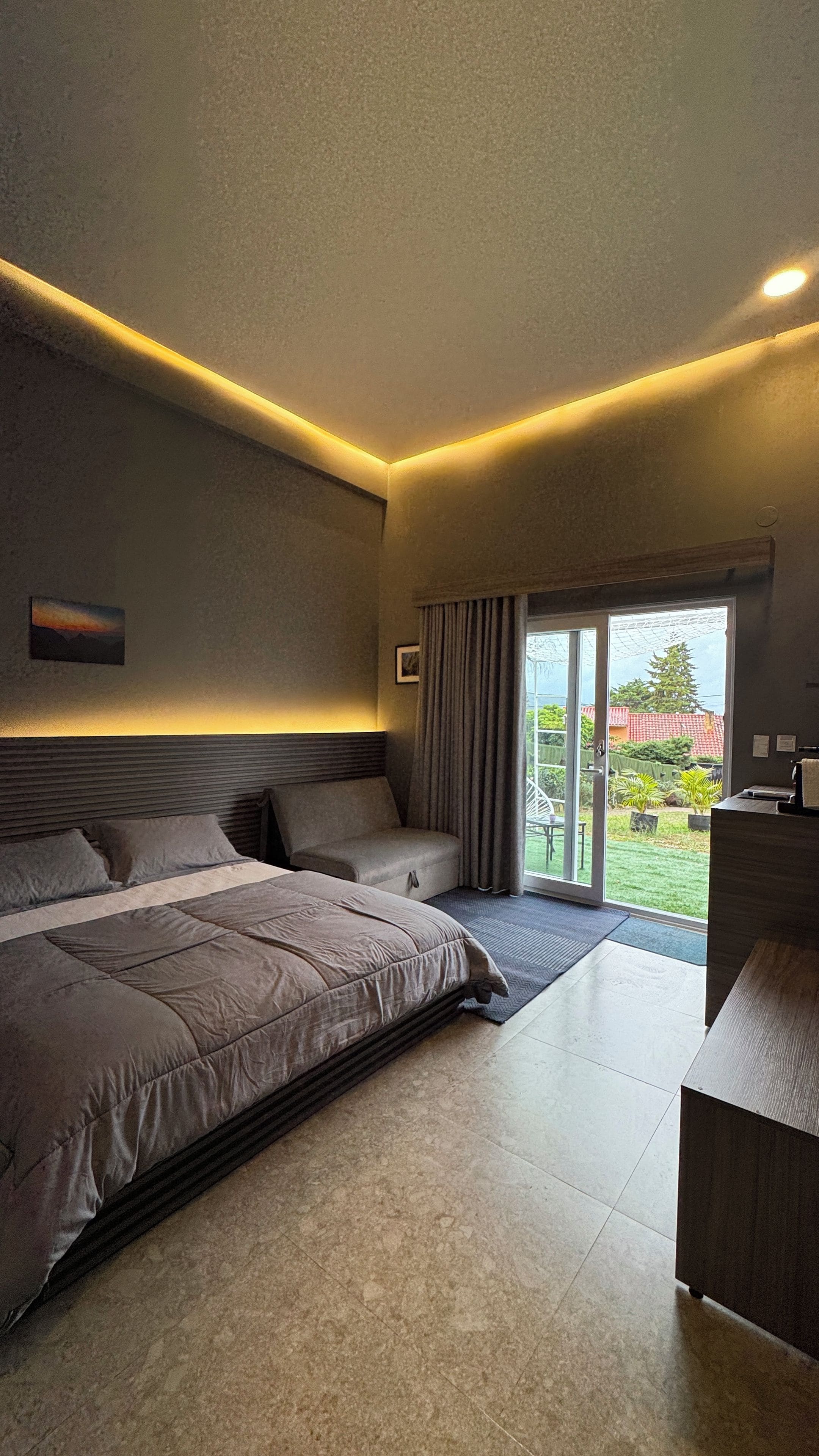 A modern bedroom with a large bed, a desk, and a view of the outdoors through a sliding glass door.