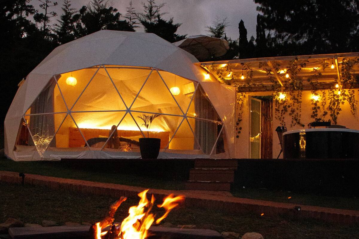 A geodesic dome structure with a fire pit in the foreground, surrounded by a garden area.