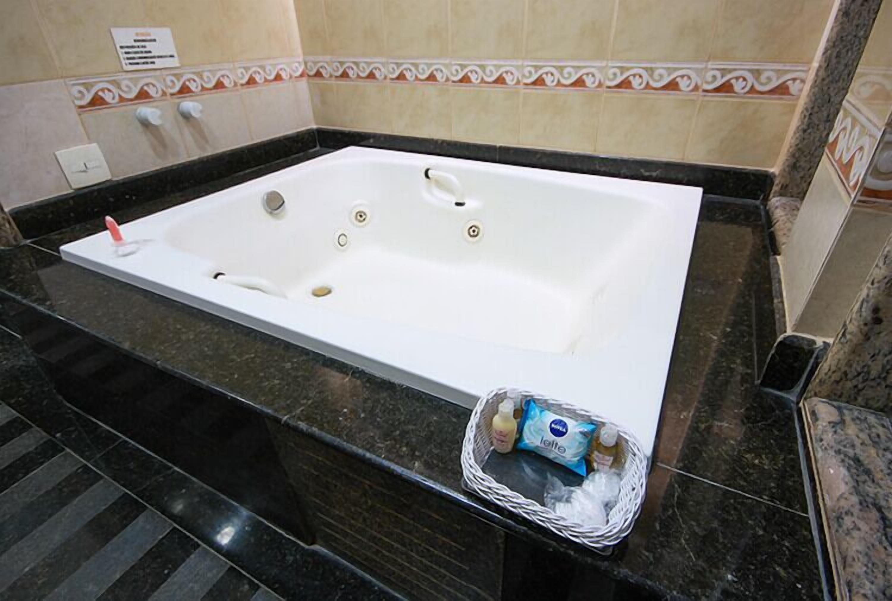 Une baignoire blanche avec une poignée, un flacon de lotion et un petit panier posés sur un comptoir en marbre noir.