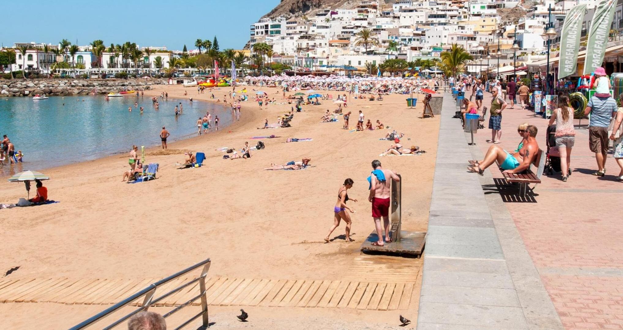 A crowded beach with people sunbathing and swimming, a wooden boardwalk, and a rocky shoreline.