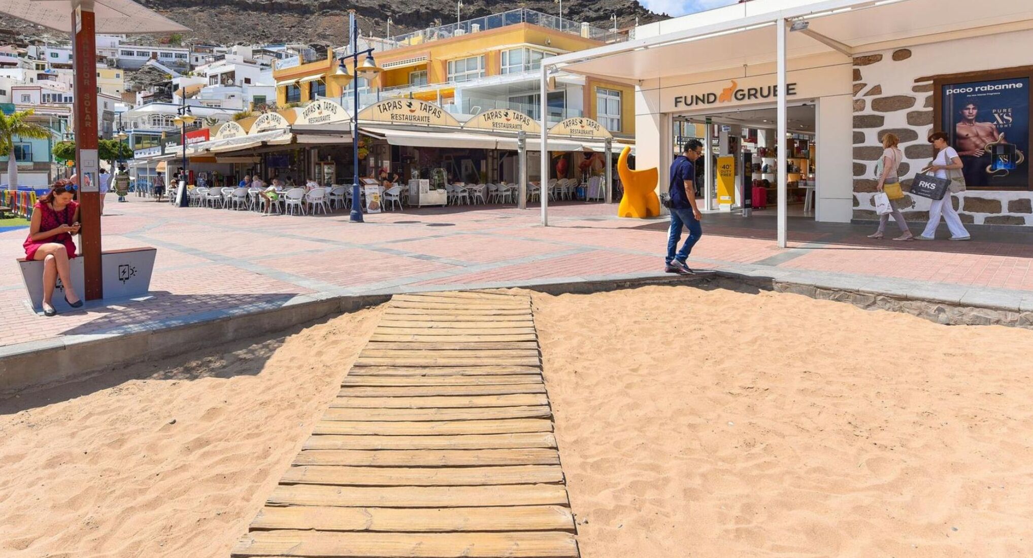 A beachside promenade with a wooden boardwalk leading to a restaurant named 'TAPAS'.