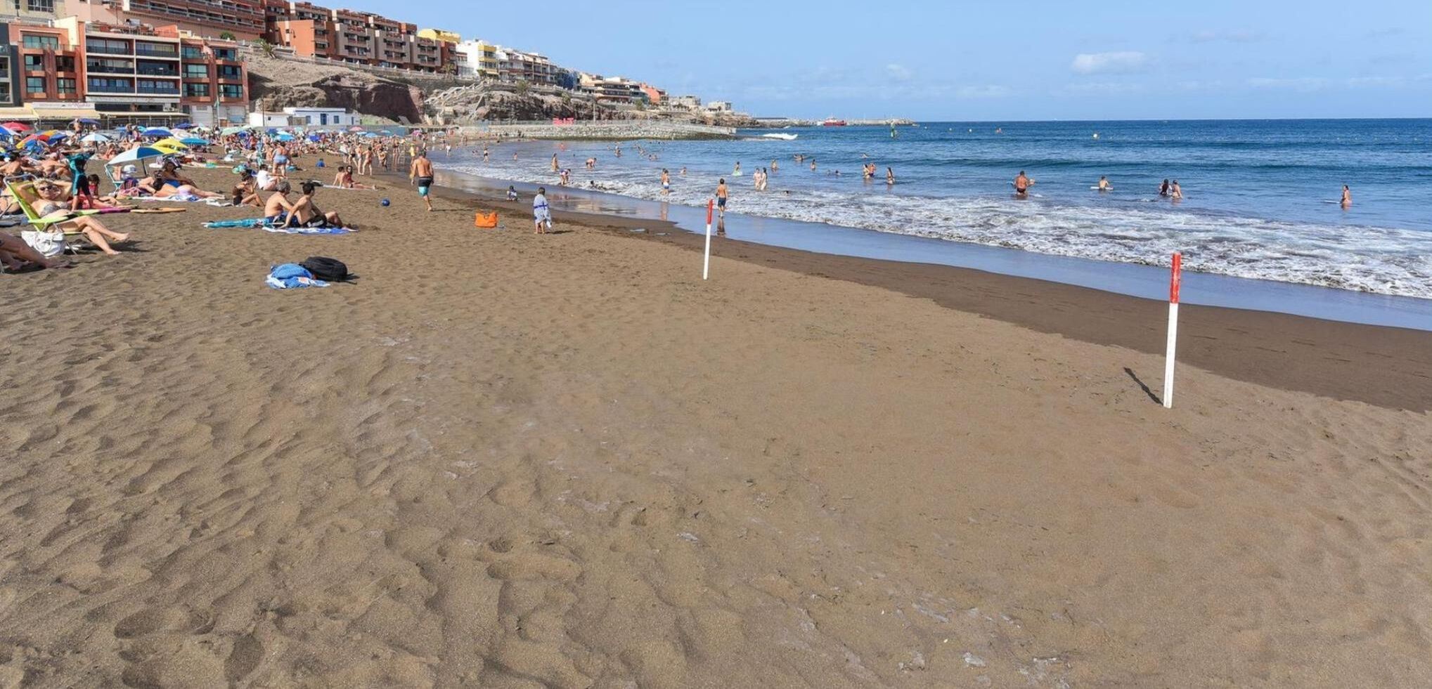 A beach with people sunbathing and swimming, buildings in the background, and a clear sky.